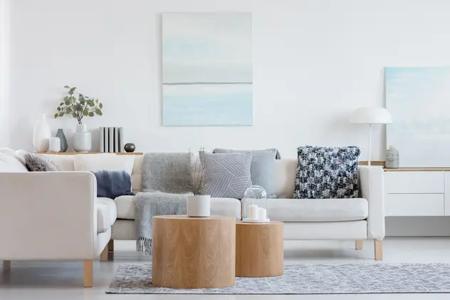 Salon w kolorze białym z jasną kanapą z drewnianymi stolikami, styl skandynawski. Element charakterystyczny: jasna kanapa z drewnianymi stolikami